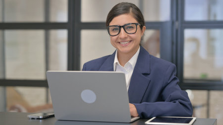 Smiling Mixed Race Businesswoman Looking at Camera while Working on Laptopの写真素材