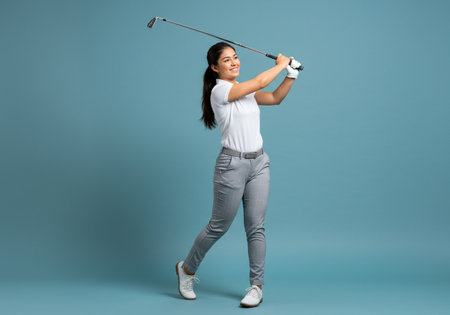 happy asian woman holding golf club and looking at camera on blue backgroundの素材