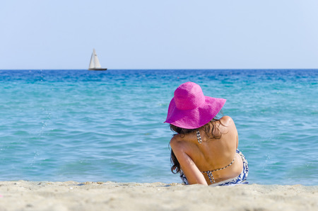 Teen straw hat girl on sea beach. Beautiful girl with pink straw hat is sunbathing on an empty beach with view of boat on blue water horizon.の写真素材