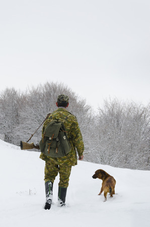 Male hunter in camouflage clothes walking on the snow field with hunting rifle during a hunt, dog follows him, foggy weatherの写真素材