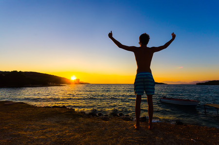 Happy celebrating winning success man at sunset or sunrise standing elated with arms raised up above his head celebratiing of having reached summit goalの写真素材