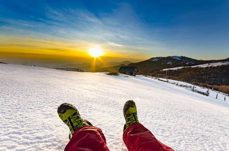 Hiker feet relaxing under the sunset lying on snow and looking at beautiful background, snow mountains and cloudsの写真素材