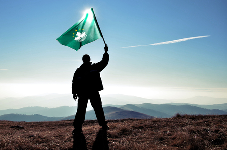 Successful silhouette man winner waving Macau flag on top of the mountain peakの写真素材