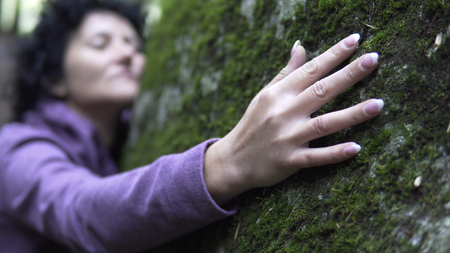 Happy woman in love relaxing hugging a huge stone with moss in forest and showing emotions. Woman is charging energy of natureの写真素材