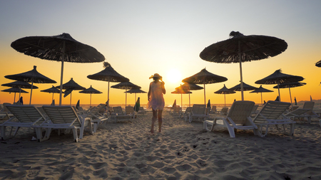 Woman in white dress and hat walk toward camera on beach in the seaside resort with sun loungers and wooden sheds against sunset, cinematicの写真素材