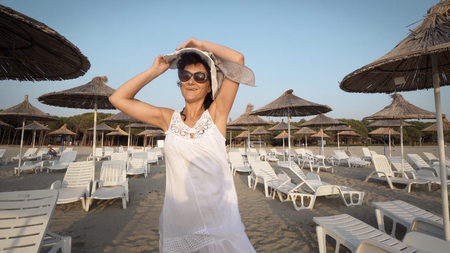 Sexy woman in white dress and hat walking on empty beautiful beach with straw umbrellas and bedsの写真素材