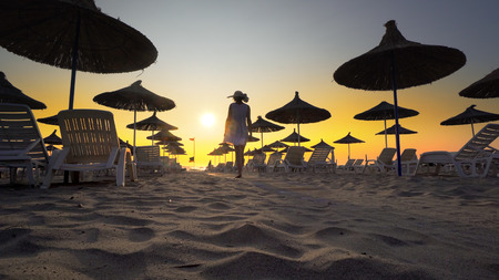 Happy woman in white dress and hat walking and turning on beach at sunset. Seaside resort with sun loungers and wooden sheds against sunset, cinematicの写真素材