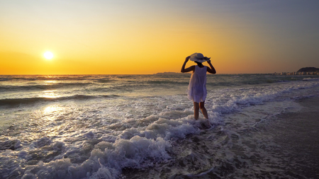 sunset scenery and woman in white dress holding hat against sunsetの写真素材