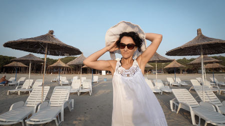 Happy smiling woman in white dress and hat blown by wind walking on empty beautiful beach with straw umbrellas and bedsの写真素材