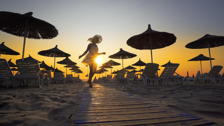 Woman in white dress and hat walk toward camera on beach in the seaside resort with sun loungers and wooden sheds against sunset, cinematicの写真素材