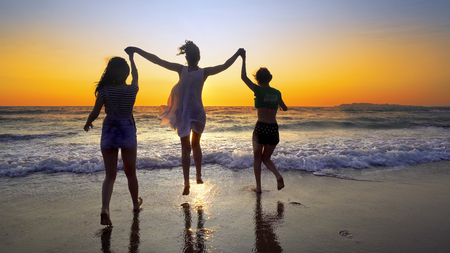 Group of people running on the beach in sunset holding for hands having fun jumping and spraying, cinematic steadicamshotの写真素材