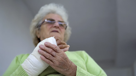 Sad old woman having pain sitting and holding her injured hand in bandagesの写真素材