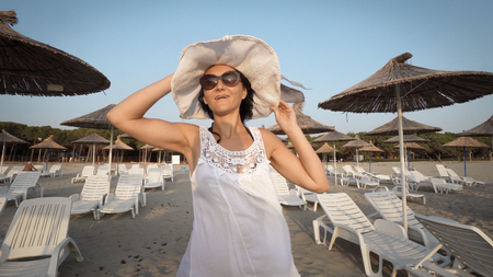 Sexy woman in white dress and hat walking on empty beautiful beach with straw umbrellas and bedsの写真素材