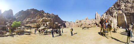 Tourists ride on camels near Royal Tombs in Petra, originally known to Nabateans as Raqmu - historical and archaeological city in Jordan. Silk and Urn Tombs on background, panoramic viewのeditorial素材