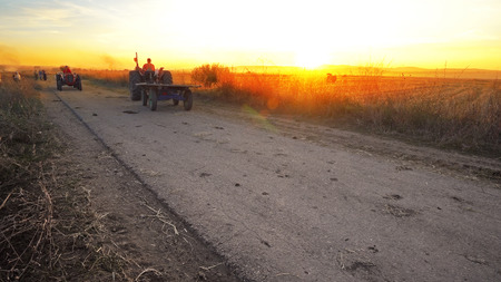 Agricultural tractor transporting harvested crops on rural road against sunset with combine harvester at backgroundの写真素材