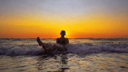 Young smiling woman sitting in sea waves and relaxing at summer vacation at sunsetの写真素材