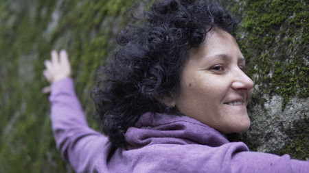 Happy woman in love relaxing hugging a huge stone with moss in forest and showing emotions. Woman is charging energy of natureの写真素材