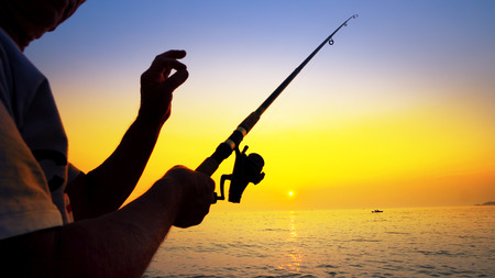 Silhouette of active fisherman fishing with rod at summer sea sunsetの写真素材