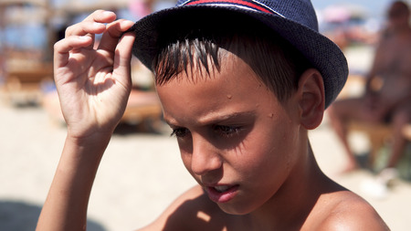 Young boy dazzled by summer sun on beach put on protection hat and smile to cameraの写真素材