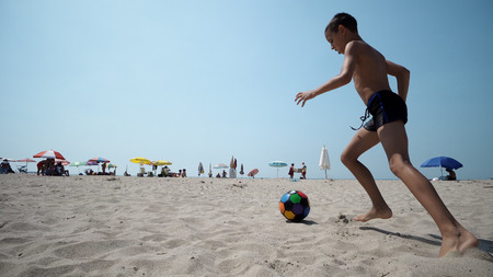 Cheerful kid playing football, running barefoot on wet sandy beach, kicking ball in slow motion, cinematic shotの写真素材