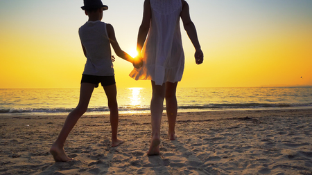 Mother with white dress and hat and son with hat standing barefoot on beach looking at the setting sun splashed by sea waves. Travel conceptの写真素材