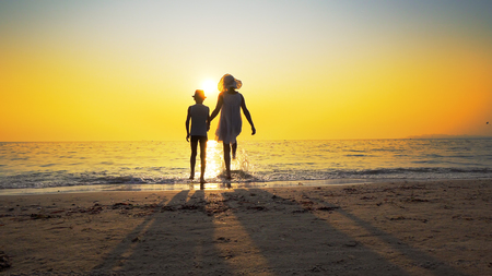 Mother with white dress and hat and son with hat standing barefoot on beach looking at the setting sun splashed by sea waves. Travel conceptの写真素材