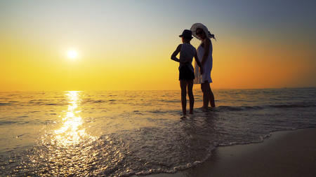 Family walk on beach to sea sunset and splash waves. Woman wear white dress and straw hat, boy has hatの写真素材