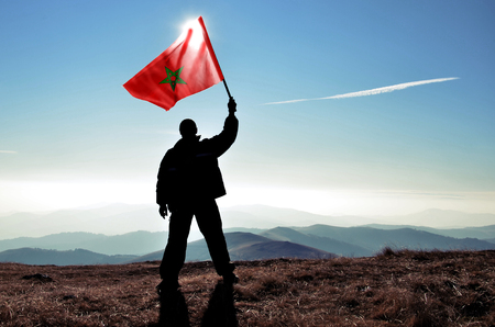 Successful silhouette man winner waving Morocco flag on top of the mountain peakの写真素材