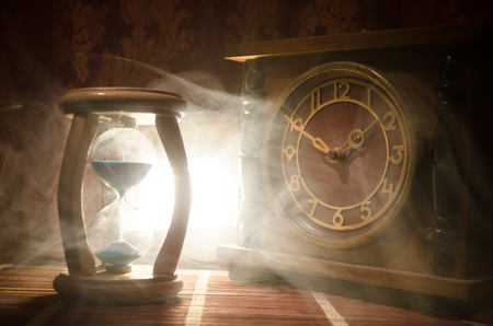 Time concept. Silhouette of Hourglass clock and old vintage wood clock with arrow and smoke on dark background with hot yellow orange red blue cold back lighting, or symbols of time with copy spaceの写真素材