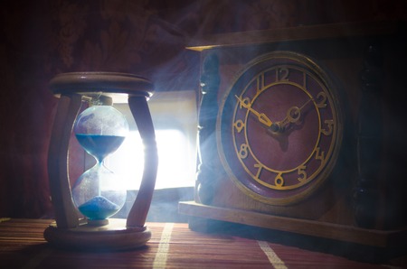 Time concept. Silhouette of Hourglass clock and old vintage wood clock with arrow and smoke on dark background with hot yellow orange red blue cold back lighting, or symbols of time with copy spaceの写真素材