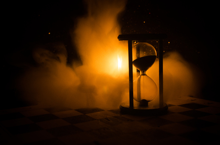 Time concept. Silhouette of Hourglass clock and smoke on dark background with hot yellow orange lighting, or symbols of time with copy space, sandglass or sand clock on chessboardの写真素材