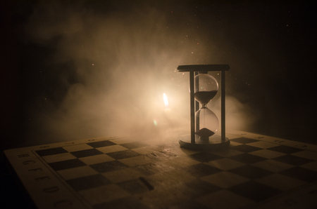 Time concept. Silhouette of Hourglass clock and smoke on dark background with hot yellow orange lighting, or symbols of time with copy space, sandglass or sand clock on chessboardの写真素材