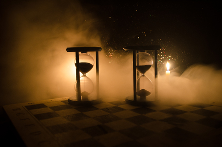 Time concept. Silhouette of Hourglass clock and smoke on dark background with hot yellow orange lighting, or symbols of time with copy space, sandglass or sand clock on chessboardの写真素材