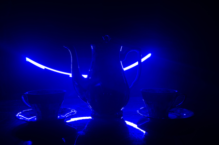 Food background tea and coffee theme. Old vintage ceramic tea or coffee pot with cups jug and sugar cup on dark background with lights and smoke. Silhouettes of ceramic tea pot and cup. Surreal decorの写真素材
