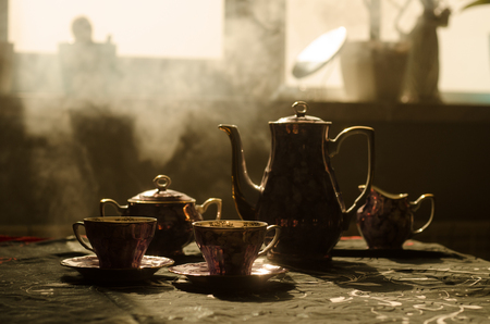 Food background tea and coffee theme. Old vintage ceramic tea or coffee pot with cups jug and sugar cup on dark background with lights and smoke. Silhouettes of ceramic tea pot and cup. Surreal decorの写真素材