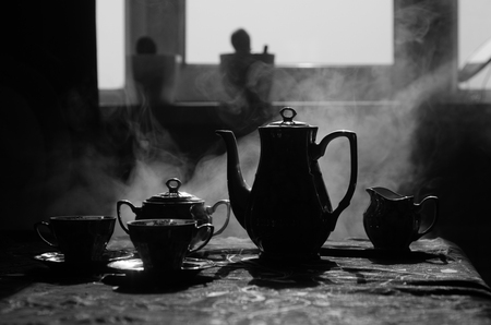 Food background tea and coffee theme. Old vintage ceramic tea or coffee pot with cups jug and sugar cup on dark background with lights and smoke. Silhouettes of ceramic tea pot and cup. Surreal decorの写真素材