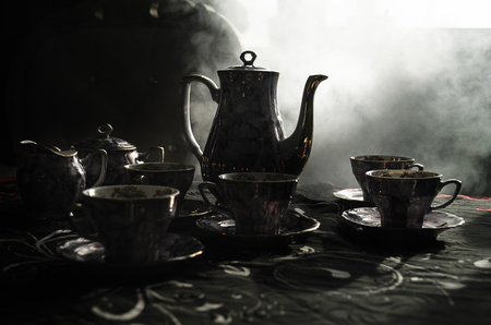 Food background tea and coffee theme. Old vintage ceramic tea or coffee pot with cups jug and sugar cup on dark background with lights and smoke. Silhouettes of ceramic tea pot and cup. Surreal decorの写真素材