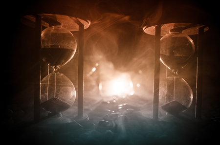 Time concept. Sand passing through the glass bulbs of an hourglass measuring the passing time as it counts down to a deadline. Silhouette of Hourglasses in smoke on dark backgroundの写真素材