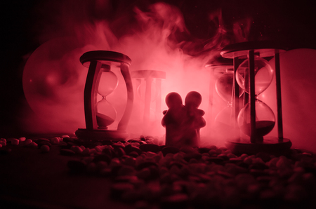 Time and love concept. Silhouettes of toy ceramic figures hugging between hourglasses in dark lighted background with fog. Couple love and time passes. Deadline of lifeの写真素材