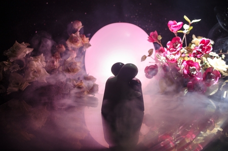 Two doll hugging on table with flowers and moon decoration Lighted background with smoke.Love concept. Greeting or gift card design idea. Silhouette of hugging coupleの写真素材