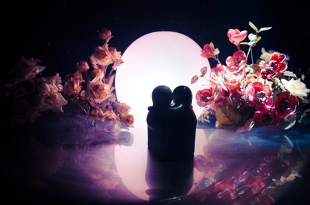 Two doll hugging on table with flowers and moon decoration Lighted background with smoke.Love concept. Greeting or gift card design idea. Silhouette of hugging coupleの写真素材