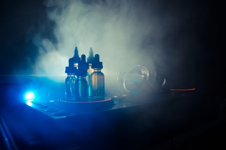 Vape and DJ Club concept. Smoke clouds and vape liquid bottles on Dj mixer close up. Light effects. Useful as background or vape or club advertisementの写真素材