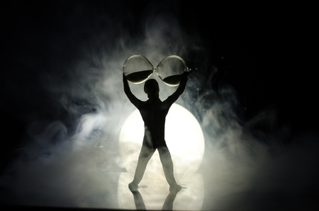 Conceptual image with sandglass and and man with moon at background with fog. Silhouette of toy figure touching hourglass. deadline concept. Man rising handsの写真素材
