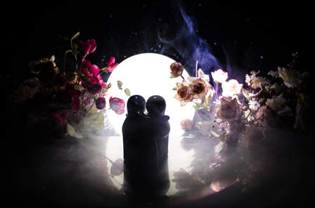 Two doll hugging on table with flowers and moon decoration Lighted background with smoke.Love concept. Greeting or gift card design idea. Silhouette of hugging coupleの写真素材