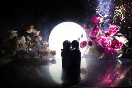 Two doll hugging on table with flowers and moon decoration Lighted background with smoke.Love concept. Greeting or gift card design idea. Silhouette of hugging coupleの写真素材