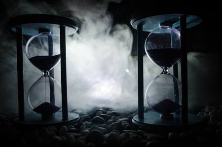 Time concept. Sand passing through the glass bulbs of an hourglass measuring the passing time as it counts down to a deadline. Silhouette of Hourglasses in smoke on dark backgroundの写真素材