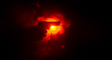 Male hand holding gun on black background with smoke ( yellow orange red white ) colored back lightsの写真素材