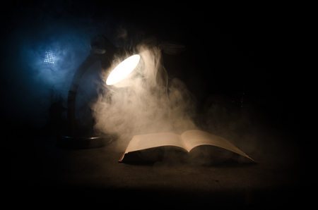 Open book near glowing table lamp on dark background, Lamp and opened book with smoke on background.の写真素材