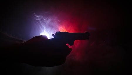 Male hand holding gun on black background with smoke ( yellow orange red white ) colored back lightsの写真素材
