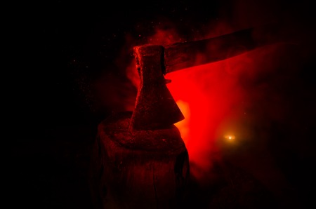 Old ax attached to the tree trunk on horror red foggy background. Scary Halloween theme with maniac killer weapon. Empty space. Selective focusの写真素材